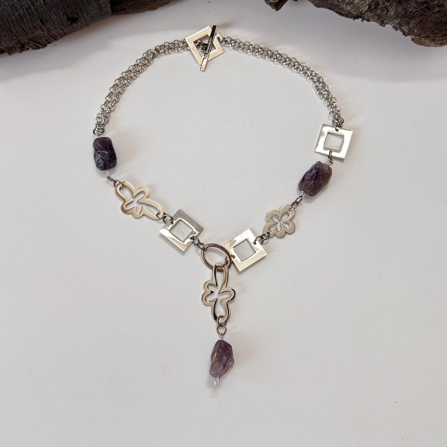 Ametist Perfection Halsband av Design by Brandeby är ett halsband i sterling silver med omväxlande geometriska och blommiga länkar med livfulla ametiststenar och en central dinglande ametist mot en vit bakgrund med träelement ovanför. Ett perfekt komplement till alla smyckens samlingar.