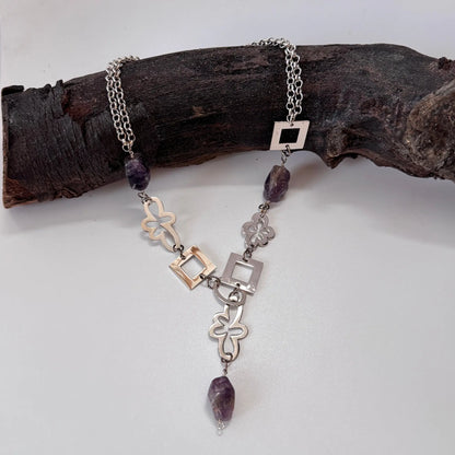 Ametist Perfection Halsband från Design by Brandeby har en kedja i sterling silver med geometriska fyrkanter och blomlänkar, accentuerad av lila stenpärlor. Den visas elegant på en mörk trägren och skapar en konstnärlig design som påminner om ett ametisthalsband.