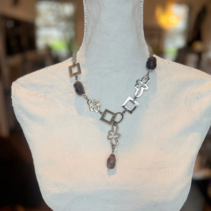 Ametist Perfection Halsband av Design by Brandeby, som visas på en vit skyltdockabyst, har ett halsband i sterling silver med geometriska former och mörka stenaccenter. Designens rutor, blommor och cirklar framkallar tidlös skönhet mot en varmt upplyst suddig bakgrund.