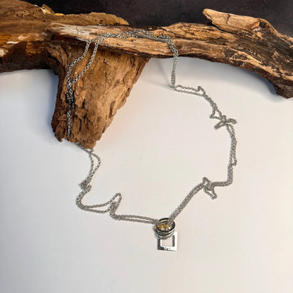 The Circle Of Life Halsband av Design by Brandeby, ett halsband med kedja i sterlingsilver med ett fyrkantigt hänge med cylinderdetalj, är elegant draperat över rustikt trä på en vit yta.