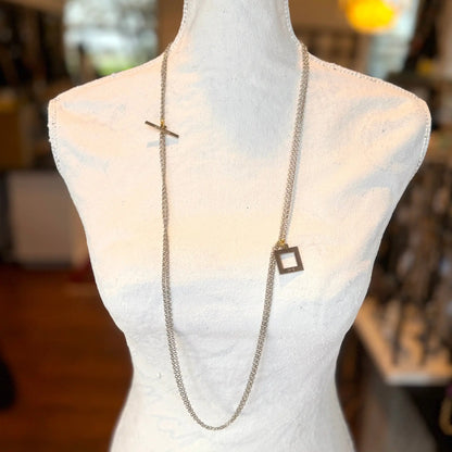 En skyltdocka visar det slående Circle Of Life Halsband av Design by Brandeby, med ett långt silverhalsband med ett T-spänne och ett fyrkantigt hänge. Bakgrunden visar ett varmt, väl upplyst rum fyllt med olika föremål som är lite oskarpa.