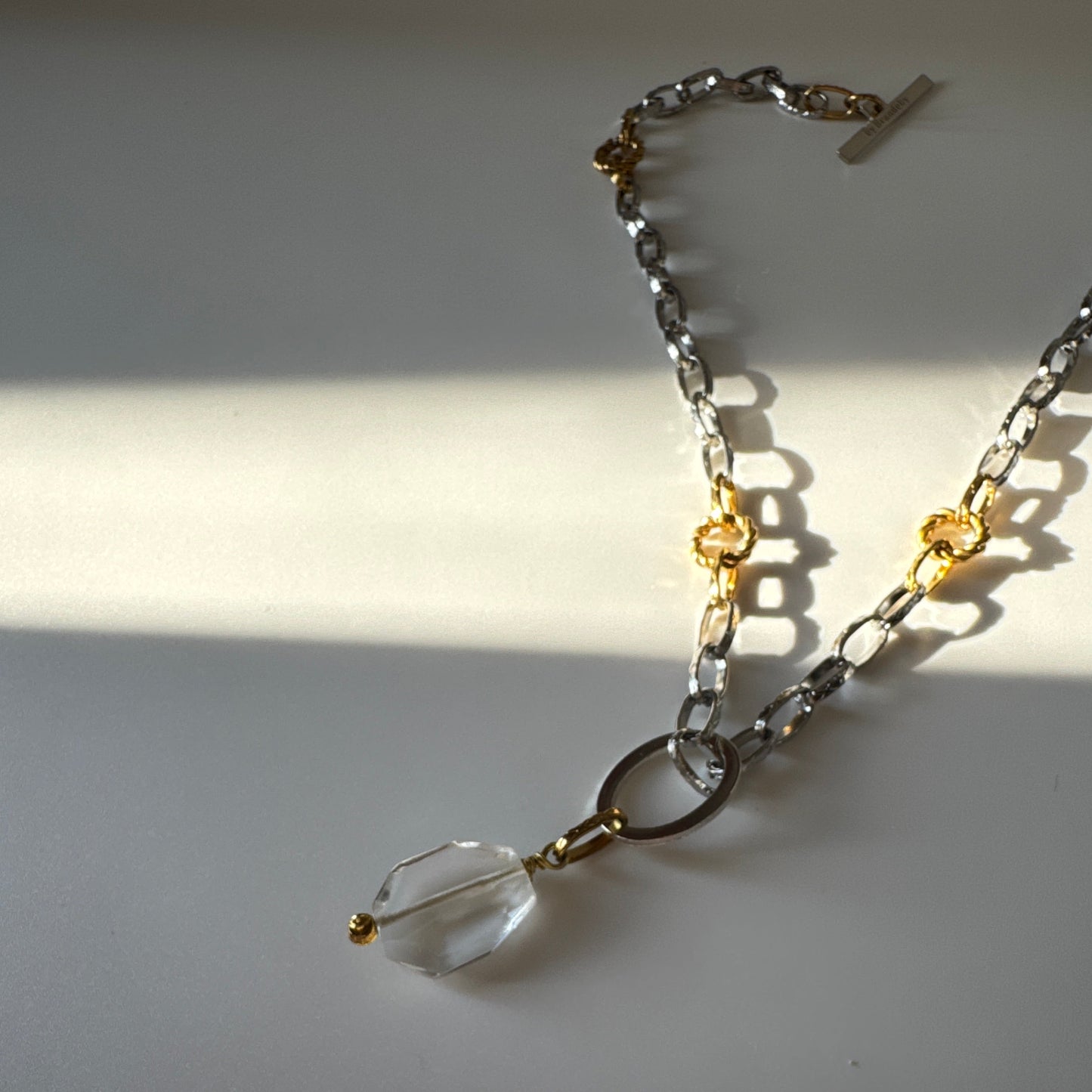 Elegant hänge med äkta bergkristall, fasade kanter och egendesignad cirkel i rostfritt stål med sterling silver-pläterade detaljer. Gulddetaljer i rostfritt stål med 18K guld PVD-beläggning. Handgjort av Design by Brandeby.